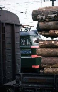 Train engine viewed through a break in a closer train, the visible part of the closer train includes a closed freight car to the left and a bit of an open flatcar with logs on the right.  