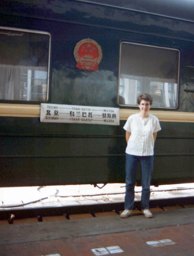 Amy (white woman; white short-sleeve shirt; jeans; short brown hair) standing in front of a train car with writing in Chinese, Russian and another language I don't know. 