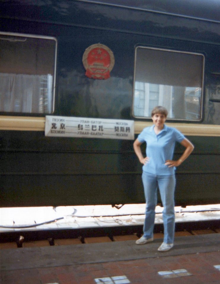 Ruth (white woman; blue t-shirt; jeans; short brown hair) standing in front of a train car with writing in Chinese, Russian and another language I don't know.