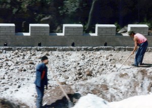 Two men in work clothes working on a portion of the top of the Great Wall that is entirely rubble.  The crenelated edge is visible in the background. 