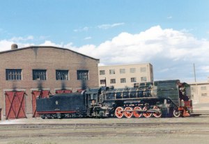 Steam freight locomotive (Chinese QJ class) with one car next to a train barn.  