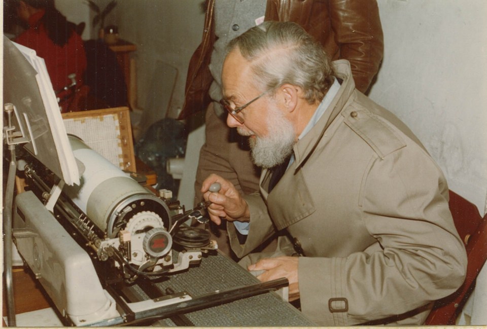 David (white man; white hair/beard; trench coat) sitting at a Chinese typewriter, poised to select a slug of type.